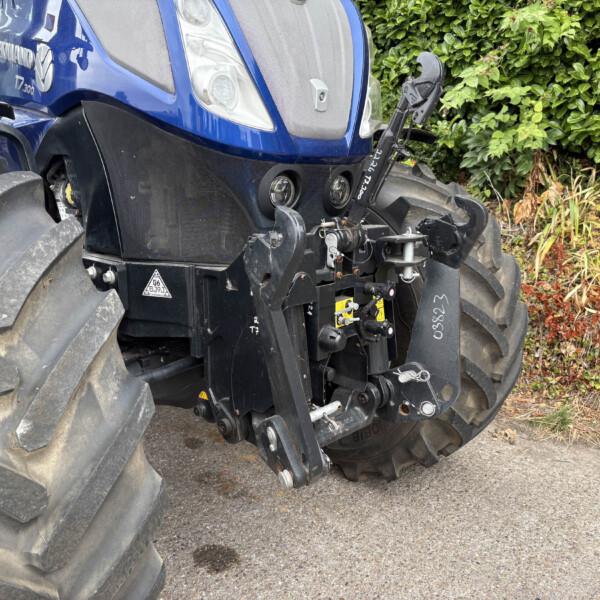 Close-up of a blue New Holland tractor's front end, showing large heavy tires, front hitch, and hydraulic components beside a green hedge.