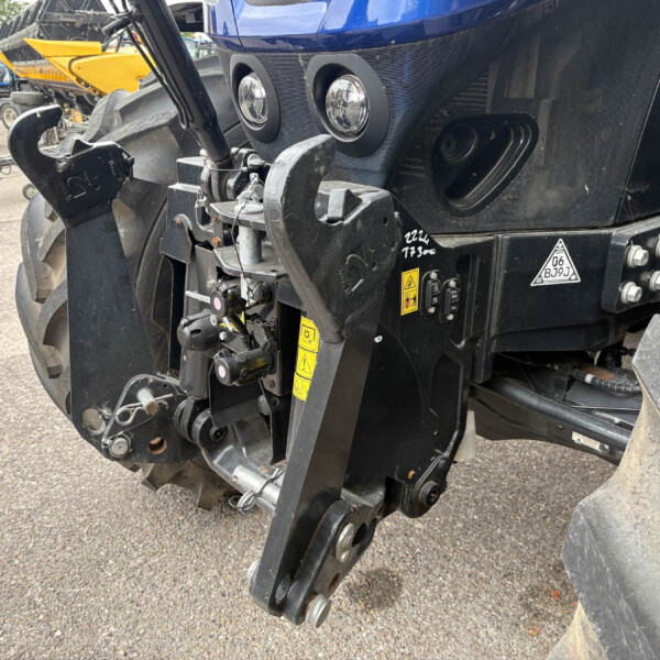 Close-up of a heavy agricultural machine's front hitch with hydraulic arms and tires nearby, showing yellow warning stickers and metal components in use.