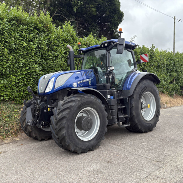 Blue New Holland tractor parked along a road beside a tall green hedge, with orange warning beacons on top of the cab.