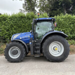 Blue New Holland T7.300 tractor parked in front of a tall green hedge under a cloudy sky.