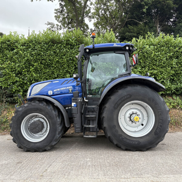 Blue New Holland T7.300 tractor parked in front of a tall green hedge under a cloudy sky.