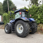 Blue New Holland tractor parked in a lot beside a tall hedge, front view with large rear tires and orange beacon on top, license plate DY24 UZR visible.