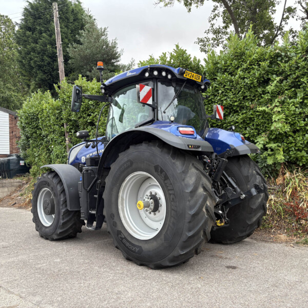 Blue New Holland tractor parked in a lot beside a tall hedge, front view with large rear tires and orange beacon on top, license plate DY24 UZR visible.