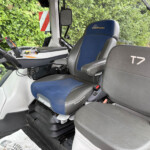 Interior view of a tractor cab with a blue-and-gray seat labeled 'Blue Power', control panel, joystick, and a mounted tablet; hedge outside the window.