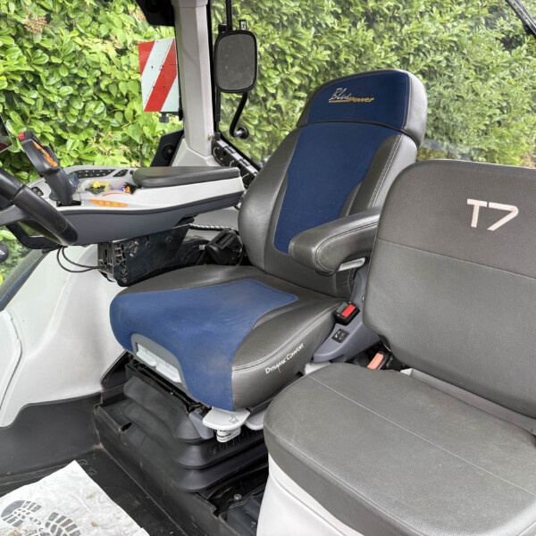 Interior view of a tractor cab with a blue-and-gray seat labeled 'Blue Power', control panel, joystick, and a mounted tablet; hedge outside the window.