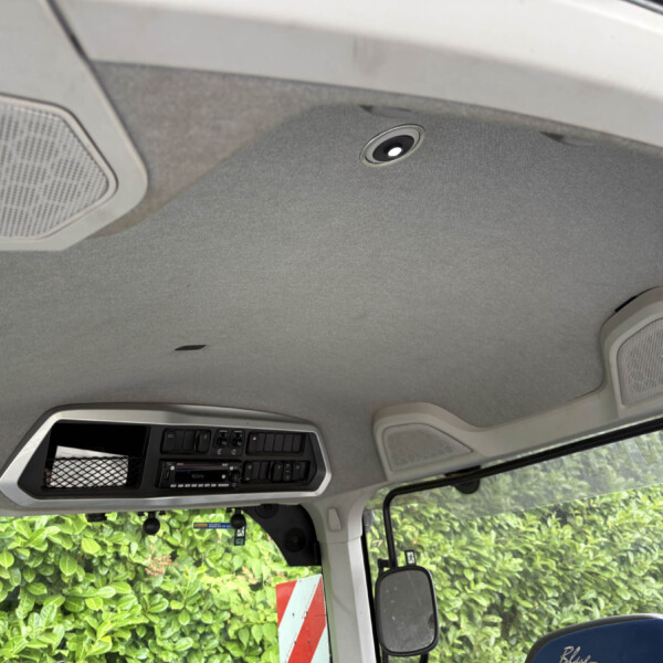Interior of a vehicle cab showing overhead console with speakers and control panel; greenery visible through side window outside the cab.