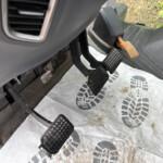 Close-up of a vehicle's driver footwell with pedals (gas, brake, and clutch) and a dirty rubber floor mat outside the pedals