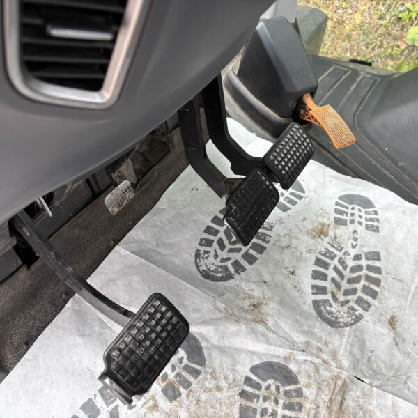 Close-up of a vehicle's driver footwell with pedals (gas, brake, and clutch) and a dirty rubber floor mat outside the pedals