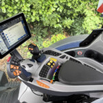 Inside a tractor cab with a mounted tablet showing configuration controls, a yellow/orange joystick, and green bushes outside the window.