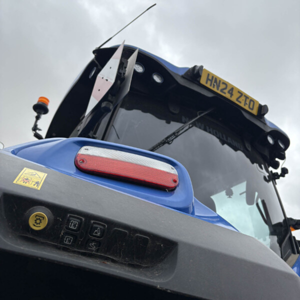 Close-up of the front of a blue tractor with an orange beacon, windshield, and license plate reading HN24 ZT0 against a gray cloudy sky.