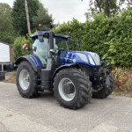 Blue New Holland T7 tractor with large rear tires parked on a paved lot, hedge and small building visible in the background.