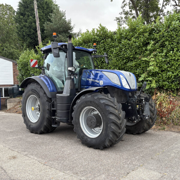 Blue New Holland T7 tractor with large rear tires parked on a paved lot, hedge and small building visible in the background.