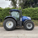 Blue New Holland tractor parked on a paved road in front of a tall green hedge, with trees in the background.