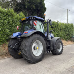 Blue New Holland tractor with oversized tires parked beside a tall green hedge on a paved road, overcast sky above.
