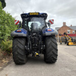 Front view of a large blue tractor with oversized rear tires, parked outdoors beside a hedge and yellow/orange agricultural equipment; license plate 'DY24 UZR' visible on top.