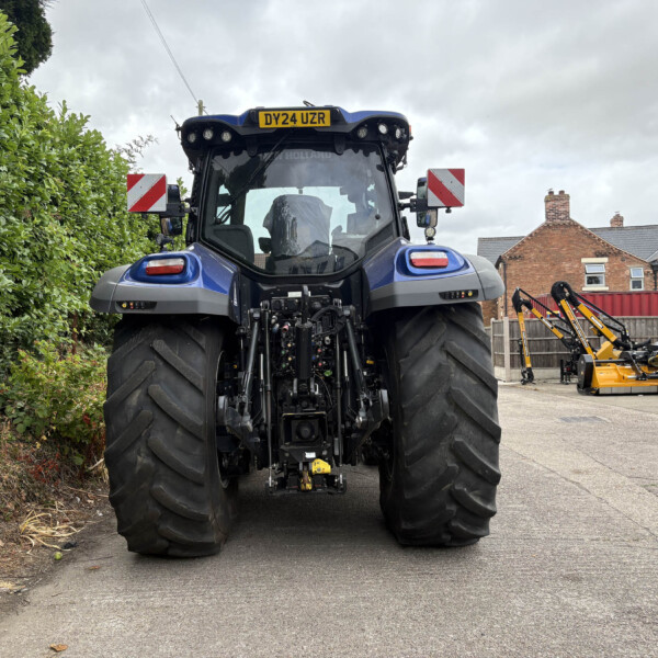 Front view of a large blue tractor with oversized rear tires, parked outdoors beside a hedge and yellow/orange agricultural equipment; license plate 'DY24 UZR' visible on top.