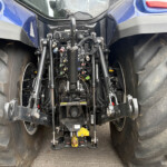 Close-up of a tractor's rear linkage with hydraulic hoses, valves, and control levers between large muddy tires between the wheels