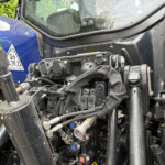 Close-up view of a blue construction vehicle cab with exposed hydraulic lines and control valves in the foreground.