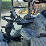 Interior of a construction vehicle cab with a large steering wheel, joystick controls, and a plastic-covered seat; exterior yellow machinery visible through the windshield
