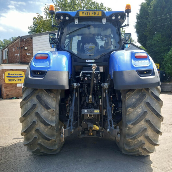 Rear view of a blue New Holland tractor with oversized rear tires, license plate YX17 FKJ, in a service yard with a yellow 'SERVICE' sign nearby