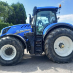 Blue New Holland tractor with large wheels parked outdoors, cabin and orange beacon visible on a sunny day.