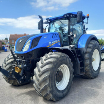 Blue New Holland tractor front view with large tires parked on a road beside a residential area under a blue sky
