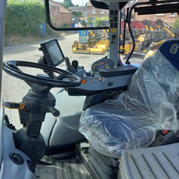 Interior of a construction vehicle cab with a large steering wheel, joystick controls, and a plastic-covered seat; exterior yellow machinery visible through the windshield
