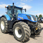 Blue New Holland tractor in the foreground with large white-rim tires, parked among other blue tractors on a sunny day.