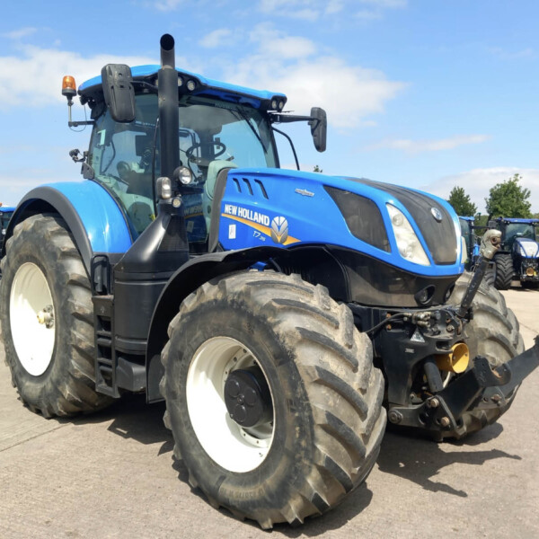 Blue New Holland tractor in the foreground with large white-rim tires, parked among other blue tractors on a sunny day.