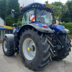 Blue New Holland tractor parked outside an agriculture shop, showing oversized tires and a yellow license plate on the cab roof.