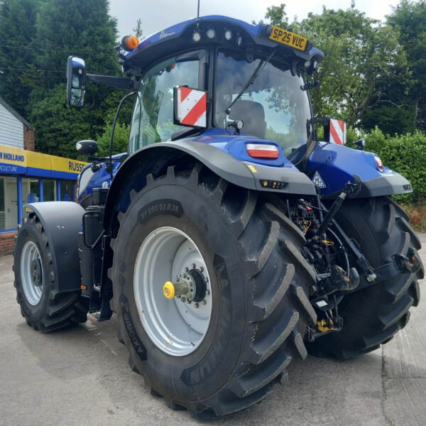 Blue New Holland tractor parked outside an agriculture shop, showing oversized tires and a yellow license plate on the cab roof.