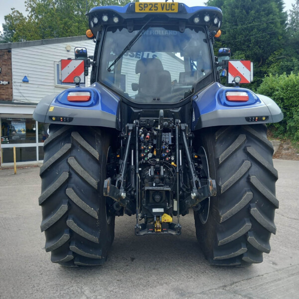 Rear view of a blue New Holland tractor with oversized tires parked outside a service shop.