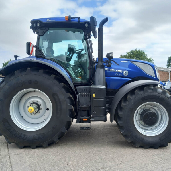 Blue New Holland tractor visible at a dealership, featuring large rear tires and a spacious cab with a service sign in the background.