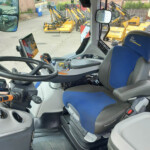 Interior of a construction vehicle cockpit showing a steering wheel, blue and gray seat, control panel, and a tablet mounted on the left; outside view reveals several yellow excavator attachments lined up.