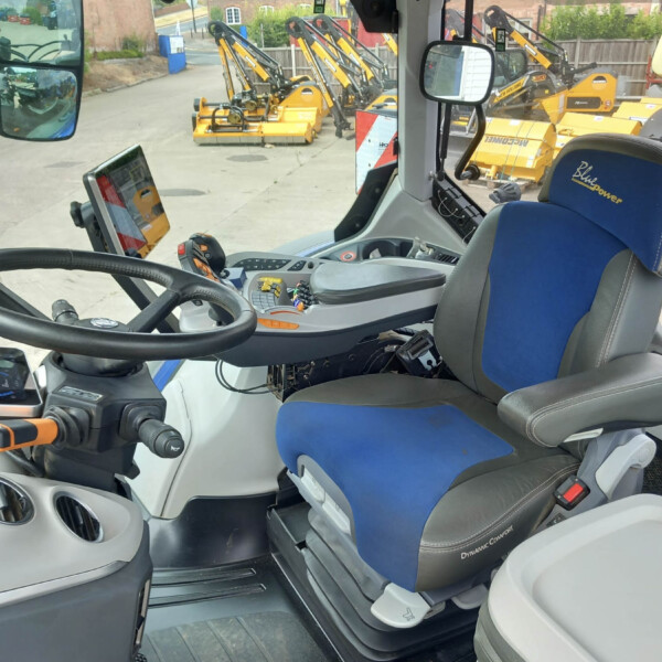Interior of a construction vehicle cockpit showing a steering wheel, blue and gray seat, control panel, and a tablet mounted on the left; outside view reveals several yellow excavator attachments lined up.