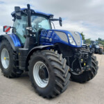 Close-up of a blue New Holland tractor with large front tires, parked in a lot with other blue tractors in the background.