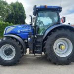 Blue New Holland T7.270 tractor with large rear tires, shown from the left-front side in a paved area with green hedges in the background and a clear cab.