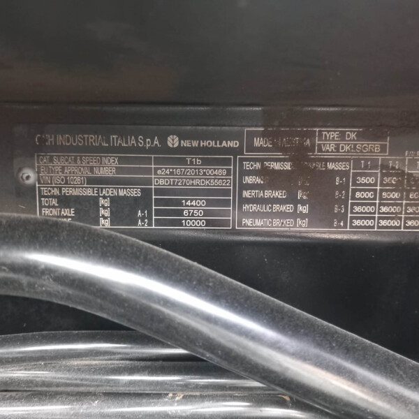 Close-up of a metal vehicle data plate under the hood showing manufacturer name (New Holland) and technical specifications such as weight and mass values (in kg).