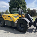 Front view of a yellow New Holland telehandler with large tires parked in a lot, with trees in the background.