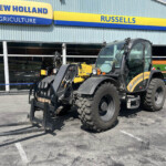 New Holland Agriculture storefront with yellow-blue sign; a large farming loader with a forklift attachment parked in front.