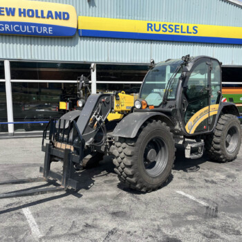 New Holland Agriculture storefront with yellow-blue sign; a large farming loader with a forklift attachment parked in front.