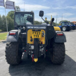 Rear view of a New Holland tractor with oversized tires, yellow rear panel and license plate FY23 TYX, lined up with blue tractors in the background at a dealership lot.