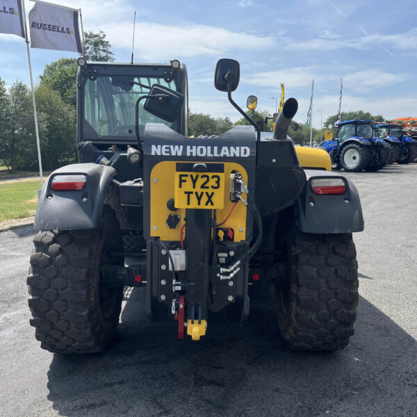 Rear view of a New Holland tractor with oversized tires, yellow rear panel and license plate FY23 TYX, lined up with blue tractors in the background at a dealership lot.