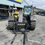 Front view of a large yellow forklift/loader with extended forks, parked in a lot by a storefront.