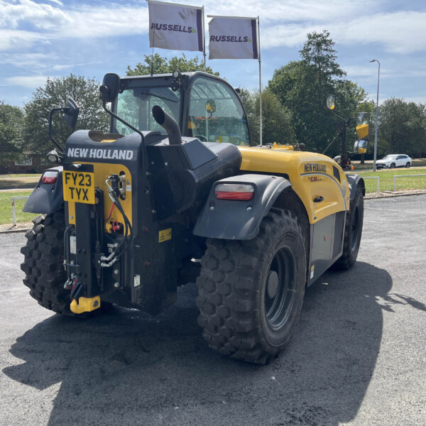 Yellow and black New Holland construction vehicle parked in a lot, with Russell flags in the background under a blue sky.