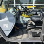 Cab interior of heavy machinery with steering wheel, levers, and a patterned seat; yellow exterior visible through window.