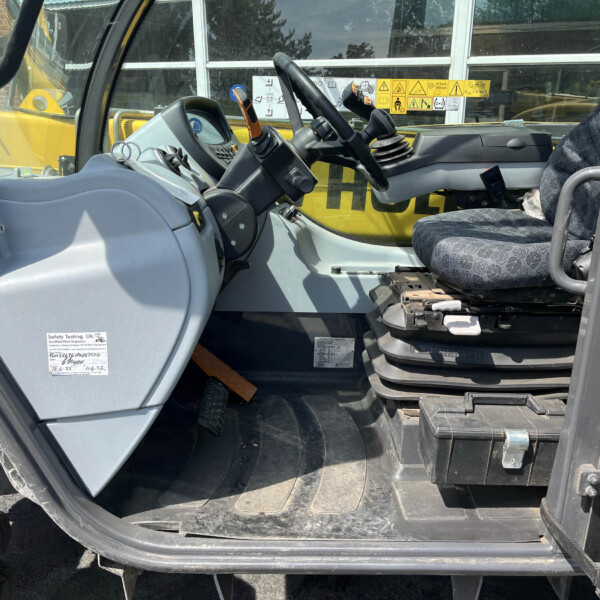 Cab interior of heavy machinery with steering wheel, levers, and a patterned seat; yellow exterior visible through window.