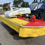 Blue tractor attached to a large white Pöttinger implement mounted on a bright yellow platform outdoors, with other farm machinery in the background.