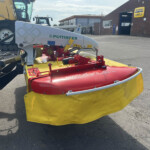 Front view of a Pottinger mower attached to a tractor in a farm equipment yard, with a service building in the background.