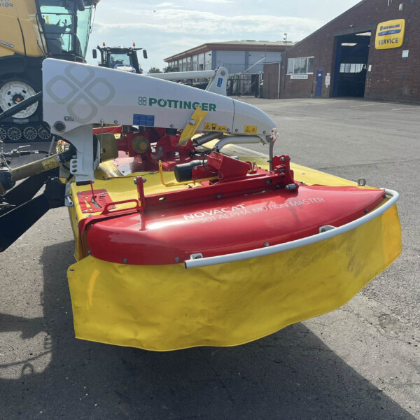 Front view of a Pottinger mower attached to a tractor in a farm equipment yard, with a service building in the background.